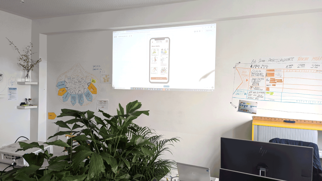 Auf einer Wand des Prozesshubs sieht man eine Beamer-Projektion mit einer App wie auch handgeschriebene Whiteboard-Skizzen